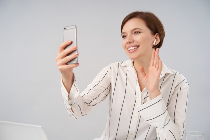 cheerful young short haired brunette woman with natural makeup raising hand hello gesture smiling pleasantly while making video call with her smartphone isolated white 700x467 - Stres Selama Work From Home? Coba Tips Berikut Ini