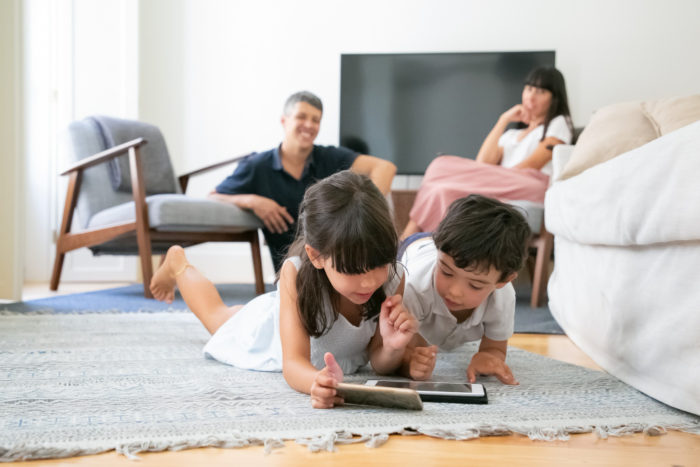 happy parent watching little kids lying floor living room using digital gadgets together 700x467 - 5 Cara Mengajar Anak Membaca di Rumah yang Menyenangkan